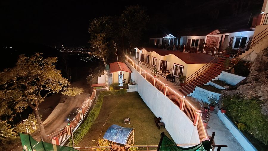 High-angle evening vista of illuminated cottages, garden lawn, and winding mountain road in Rishikesh