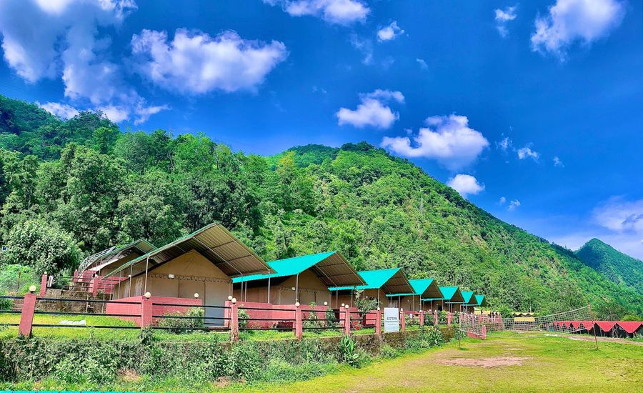 Row of safari-style camping huts set against lush green hills under a bright blue sky in Rishikesh