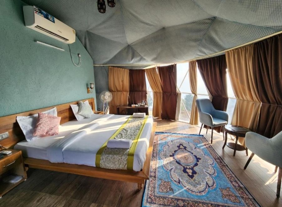 Sunlit interior of luxury glamping dome featuring plush double bed, armchairs, and scenic wrap-around windows in Rishikesh